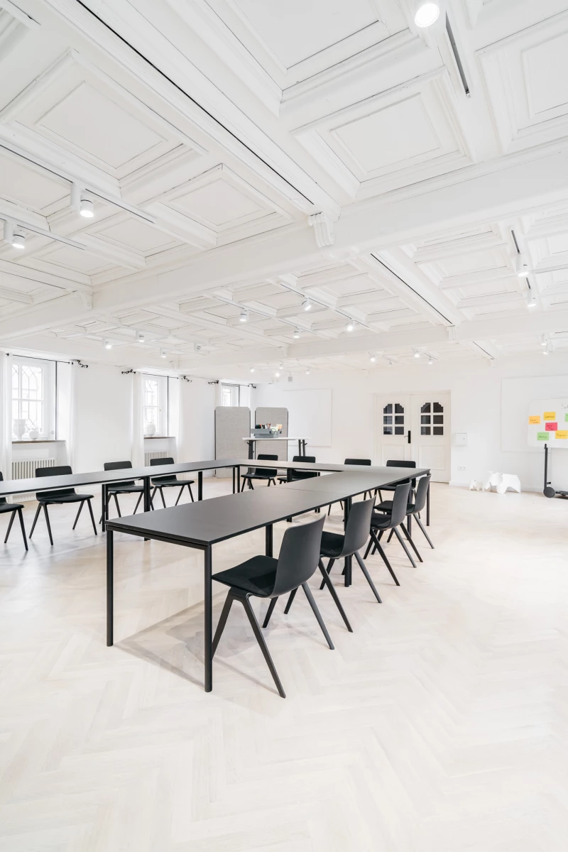 Heller, minimalistischer Schulungsraum mit großen Tischen und schwarzen Stühlen. Weiße Decke mit Holzbalken und große Fenster sorgen für eine offene, moderne Atmosphäre. Die Einrichtung wirkt schlicht und funktional, ideal für Meetings oder Seminare.