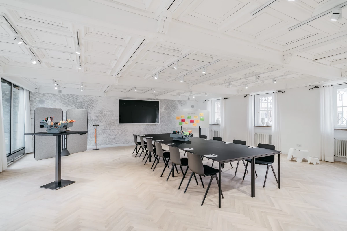 Moderner, heller Besprechungsraum mit großem schwarzem Tisch und schwarzen Stühlen. Weiße Decke mit Einbauleuchten, große Fenster und eine Pinnwand mit bunten Haftnotizen an der Wand. Ein multifunktionaler Raum für Meetings und Workshops.