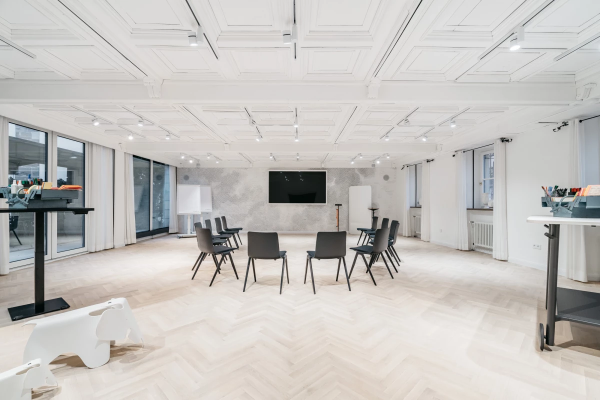 Moderner, heller Schulungsraum mit weißen Wänden und Decke. Mehrere schwarze Stühle sind in einem Halbkreis angeordnet, an der Wand hängt ein großer schwarzer Bildschirm. Große Fenster lassen viel Tageslicht herein und verleihen dem Raum eine offene, einladende Atmosphäre.
