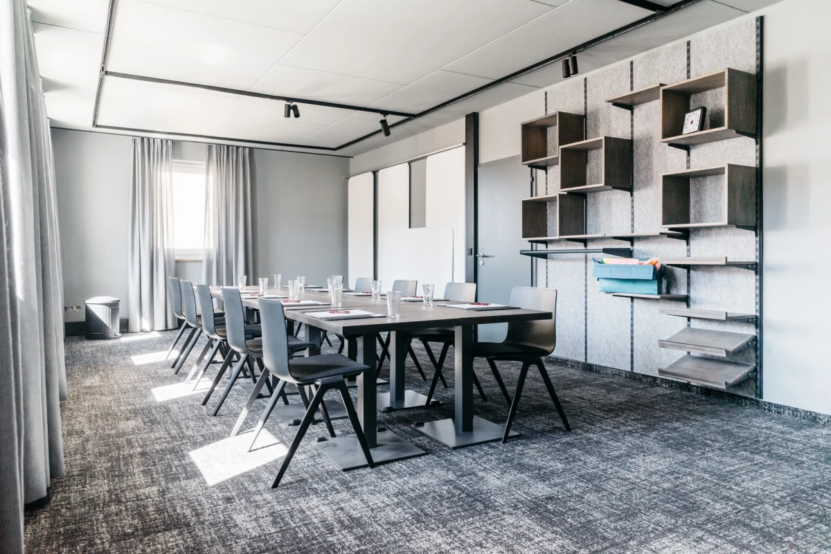 Moderner Konferenzraum mit großem schwarzem Tisch, grauen Stühlen und strukturierter Teppichoberfläche. An der Wand offene Regale und Wandschränke in gedeckten Farben, große Fenster mit hellen Vorhängen lassen natürliches Licht herein.