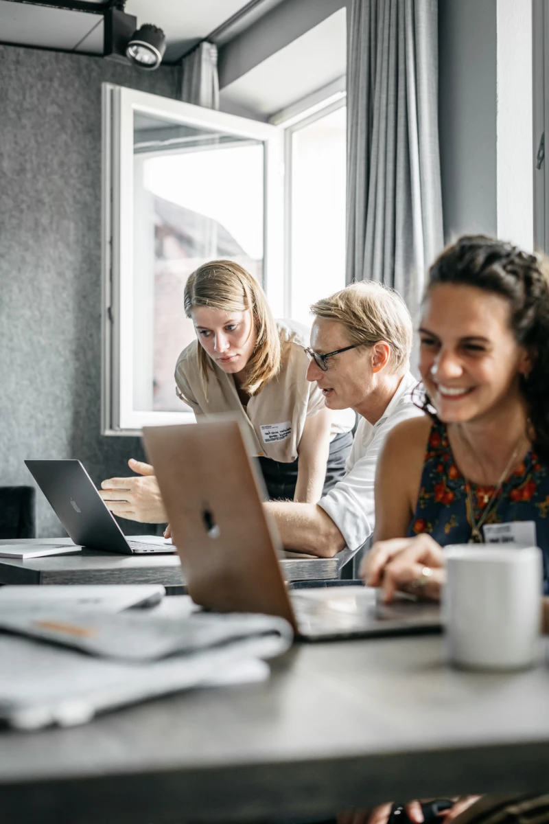 Drei Personen arbeiten konzentriert an Laptops an einem Tisch nahe einem Fenster. Die Stimmung ist entspannt und produktiv, mit einer Kaffeetasse im Vordergrund. Sie scheinen in einem modernen Büro oder Besprechungsraum zu sein.