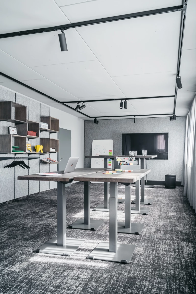 Moderner Büroraum mit höhenverstellbaren Schreibtischen, grauen Bodenfliesen und weißen Wandregalen. Schwarze Deckenspots erhellen den minimalistischen Arbeitsbereich mit offenem, hellem Raumkonzept.