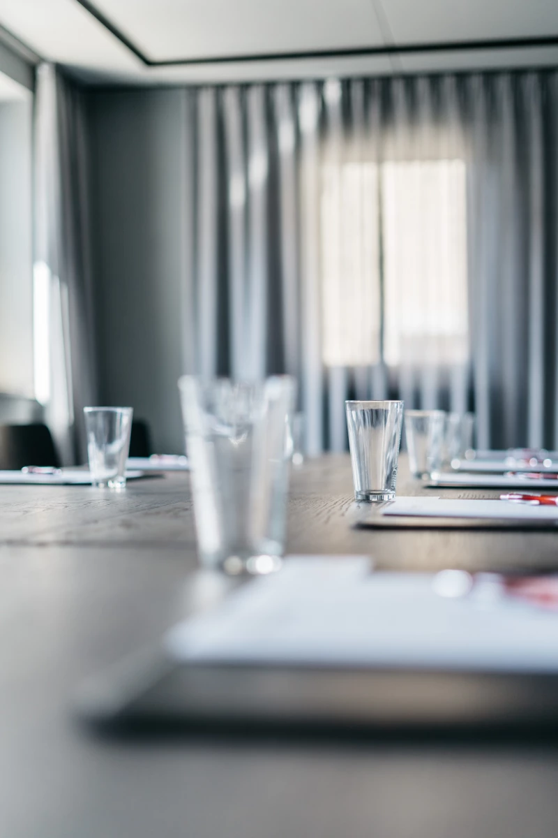 Konferenztisch mit Wassergläsern und Dokumenten, umgeben von gedämpften grauen Vorhängen und einem unscharfen Fenster im Hintergrund. Professionelle Meetingatmosphäre in einem ruhigen, minimalistischen Besprechungsraum.