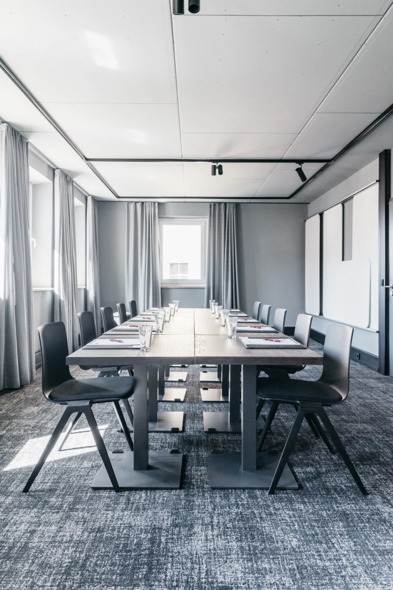 Moderner Konferenzraum mit langem Holztisch und grauen Bürostühlen, umgeben von weißen Wänden und bodenlangen Vorhängen. Tageslicht fällt durch die Fenster und erhellt den minimalistischen, professionellen Raum.