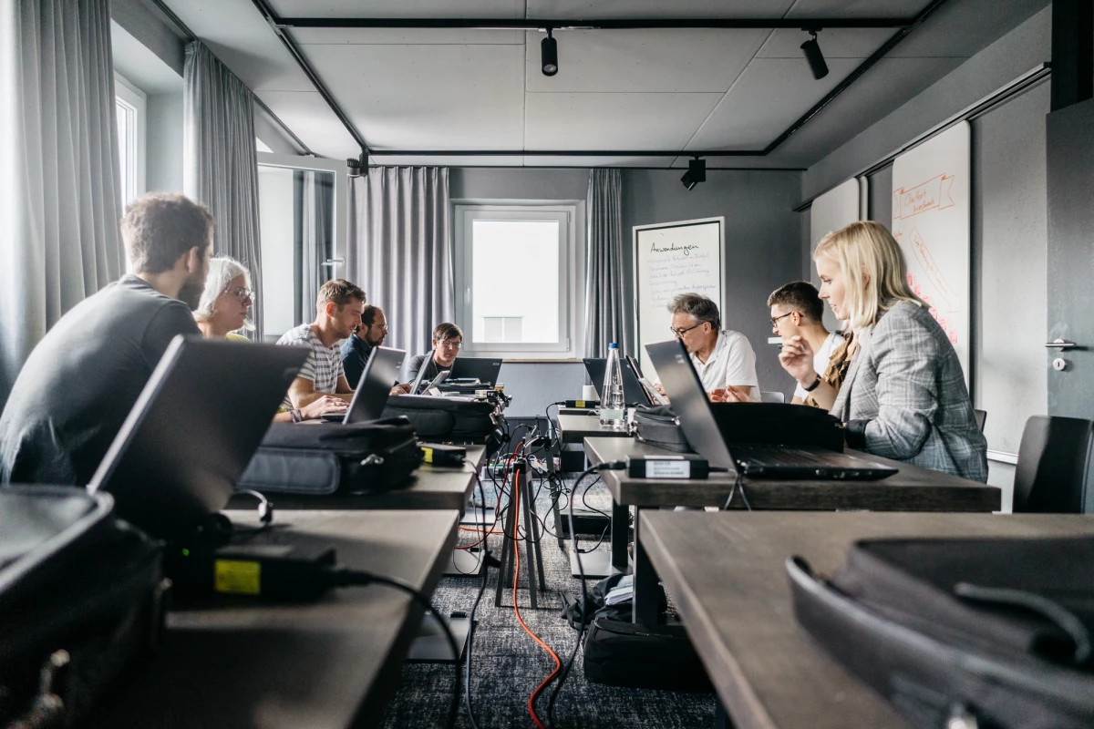 Mehrere Personen arbeiten konzentriert an Laptops in einem modernen Besprechungsraum mit grauen Vorhängen und Fenstern. Die Teilnehmer sitzen an einem langen Tisch, auf dem Laptops, Kabel und technische Geräte verteilt sind. Die Atmosphäre wirkt professionell und fokussiert.
