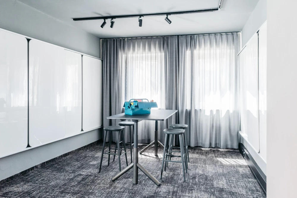 Moderner Konferenzraum mit grauen Vorhängen und Teppichboden. Hochstehtische mit Barhockern und eine blaue Tasche stehen bereit. Schwarze Schienenstrahler an der Decke sorgen für optimale Beleuchtung.