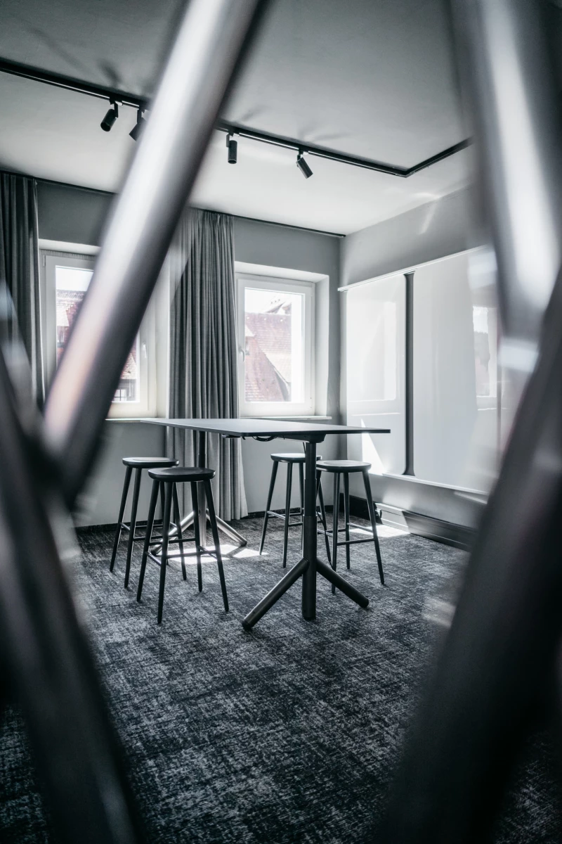 Moderner Besprechungsraum mit hohen Bartischen und Hockern auf grauem Teppichboden. Große Fenster mit Vorhängen und Deckenspots sorgen für helle, elegante Atmosphäre. Blick durch einen verschwommenen Vordergrund, der die Tiefe des Raumes betont.