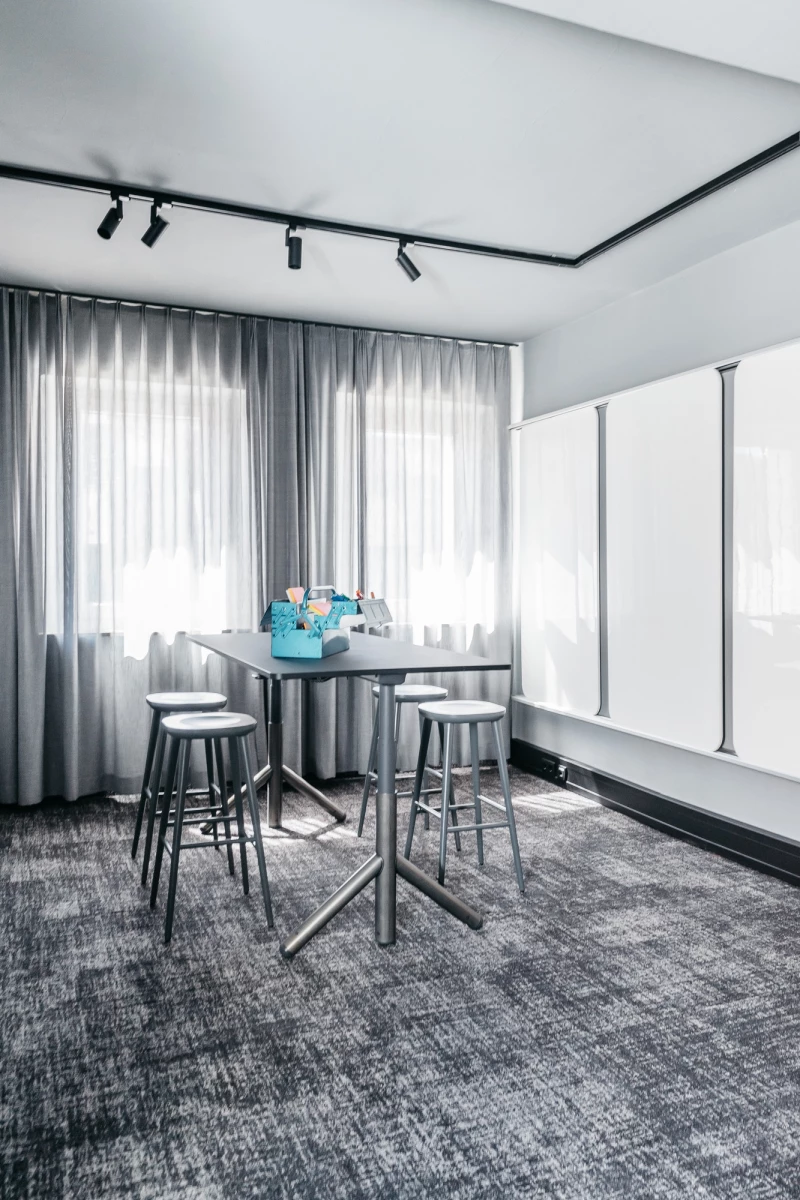 Kleiner Meetingraum mit grauem Teppichboden, einem runden Stehtisch und drei Barhockern. Große Fenster mit weichen, hellen Vorhängen und Deckenspots sorgen für eine helle, moderne Atmosphäre.