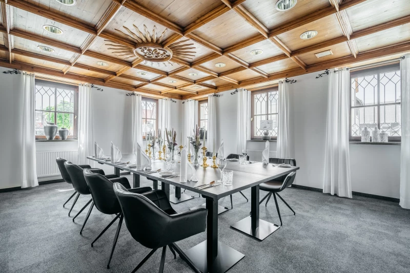 Moderner Konferenzraum mit großen Fenstern und Holzdecke. Langer weißer Tisch mit schwarzen Designerstühlen, dekoriert mit Vasen und Kerzen. Helle, elegante Atmosphäre mit natürlichem Tageslicht.