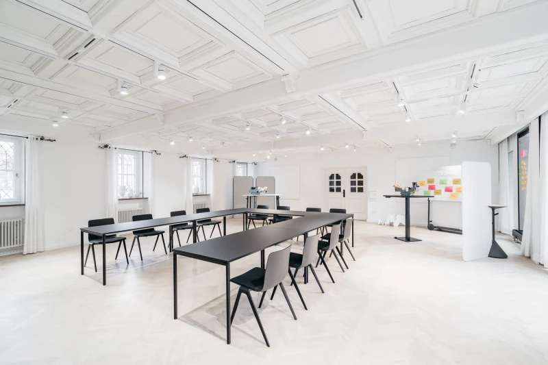 Heller, minimalistischer Seminarraum mit langen schwarzen Tischen und passenden Stühlen. Weiße Wände und Decke mit Oberlicht, große Fenster sorgen für natürliches Licht. An der Wand hängen bukte Haftnotizen, ein Whiteboard steht bereit für Besprechungen und Workshops.