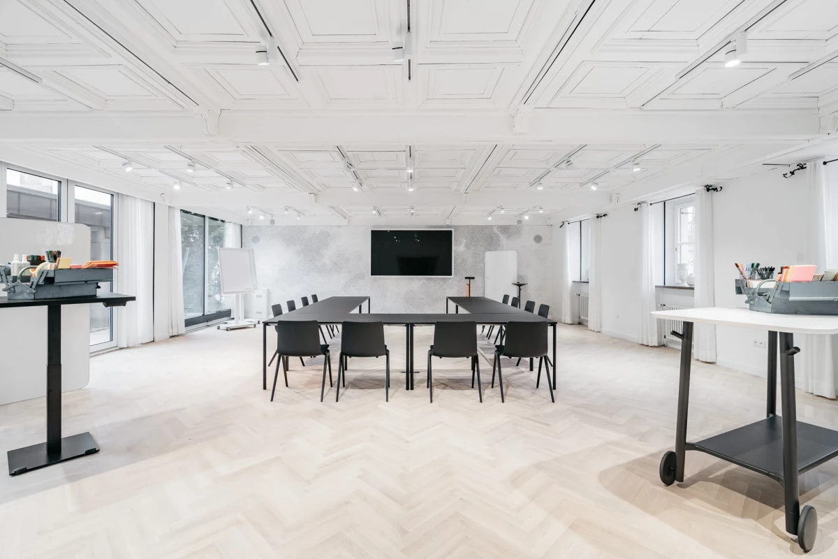 Moderner, heller Konferenzraum mit großer schwarzer Konferenztafel und mehreren schwarzen Stühlen um einen Tisch. Weiße Wände, große Fenster und ein heller Holzboden prägen den minimalistischen, professionellen Meetingraum.