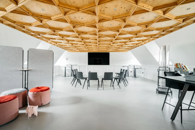 Moderner Konferenzraum mit geometrischer Holzdecke, grauen Stühlen um einen Tisch und roten Polstersesseln. Ein schwarzer Bildschirm hängt an der Wand, und der Raum hat eine helle, minimalistisch gestaltete Atmosphäre.