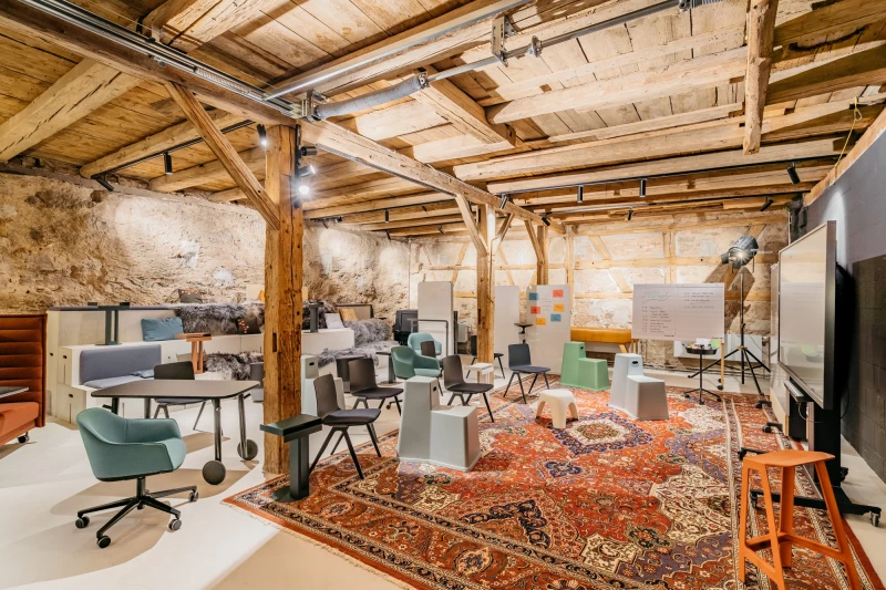 Rustikaler Konferenzraum mit Holzbalkendecke und -ständern, ausgestattet mit modernen Bürostühlen und einem großen orangefarbenen Vintage-Teppich. Industrieller Charme trifft auf zeitgenössisches Design in einem umgebauten Scheunenloft mit Steinwänden und hellen Möbeln.