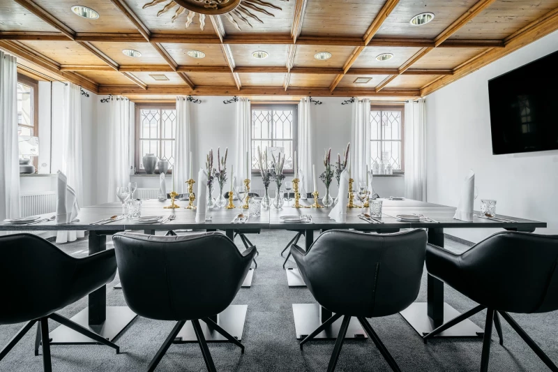 Eleganter Konferenzraum mit großen Fenstern, weißer Wandgestaltung und holzgetäfelter Decke. Langer Tisch mit schwarzen Drehstühlen und festlicher Tischdekoration mit goldenen Kerzenhaltern. Modernes, helles Ambiente für Meetings und Veranstaltungen.