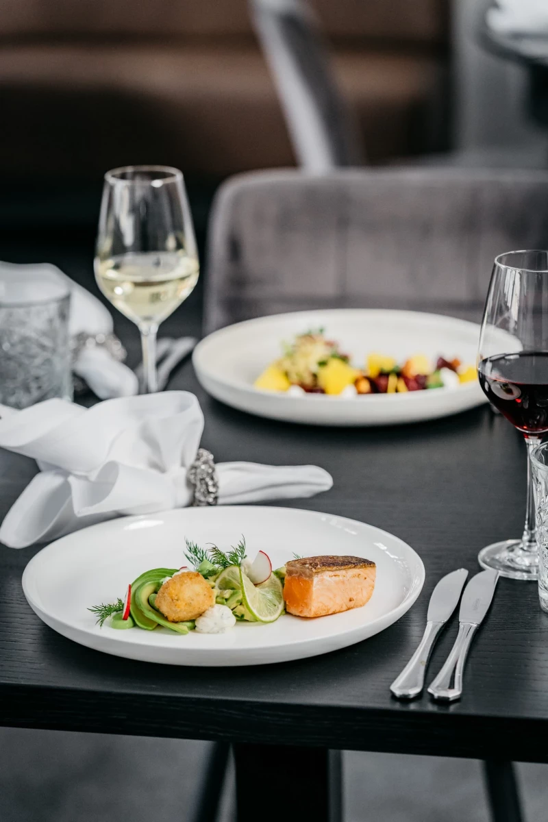 Elegante Restaurantteller mit zartem Lachsfilet, begleitet von einem Weißweinglas und einem Rotwein. Fein angerichtet auf einem dunklen Tisch mit weißer Serviette und Besteck, eine geschmackvolle Komposition aus Gourmet-Küche.