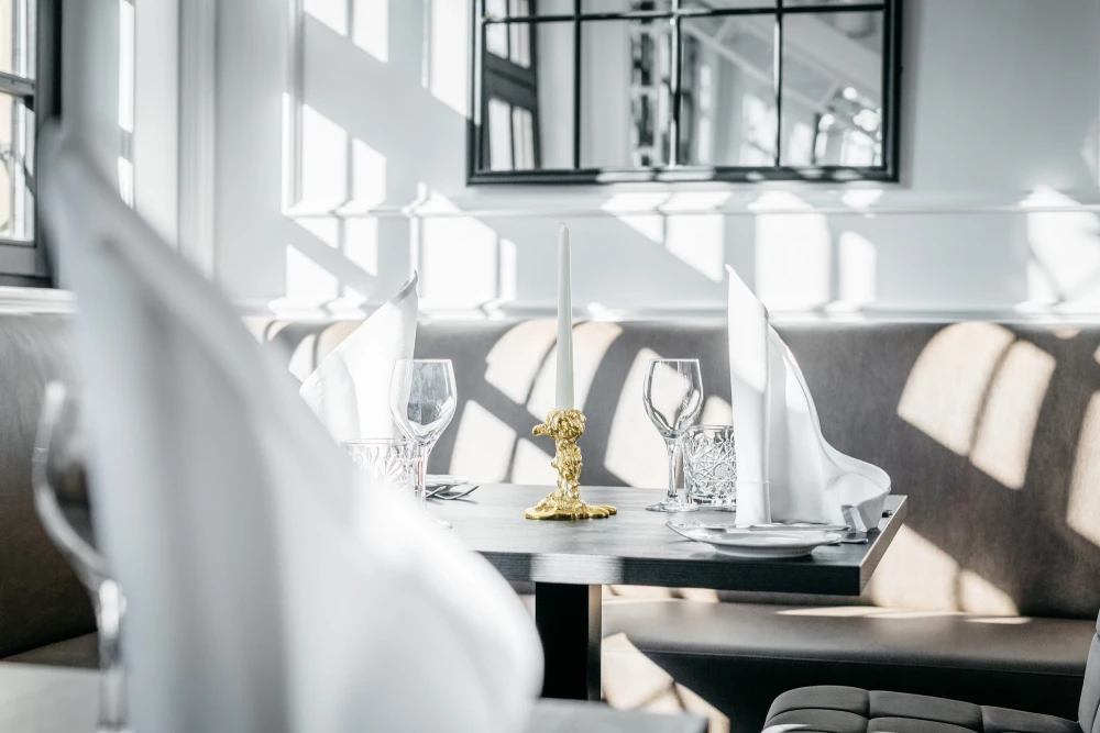 Elegante Tischdekoration in einem hellen, modernen Restaurant-Ambiente mit gedeckten Tischen, weißen Stühlen und großen Fenstern, die weiches Tageslicht hereinlassen.