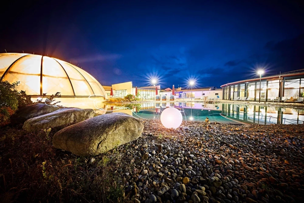 Nächtliche Ansicht der Frankentherme mit beleuchtetem Außenbecken, Kieselsteinen im Vordergrund und einer glühenden Kugel, umgeben von modernen Gebäuden mit Lichtern und blauem Abendhimmel.