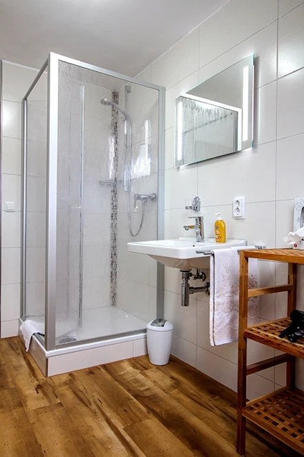 Modernes, helles Badezimmer mit Duschkabine aus Glas, weißen Fliesen und Holzboden. Holzregal mit Handtuch, weißes Waschbecken und Spiegel an der Wand, gelbe Duschutensilien auf der Ablage.
