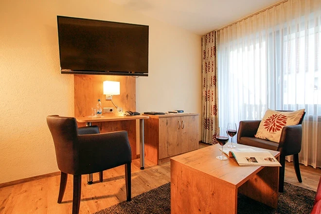 Hotelzimmer mit Holzmöbeln, Fernseher an der Wand und einem Schreibtisch mit Bürostuhl. Ein Sessel mit gemustertem Kissen steht neben einem Couchtisch mit Weinglas und Zeitschrift, große Fenster mit hellen Vorhängen lassen natürliches Licht herein.