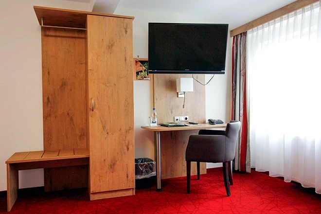 Hotelzimmer mit rotem Teppich, Holzschrank, Schreibtisch mit schwarzem Stuhl und an der Wand montiertem Flach-TV. Helle Gardinen lassen natürliches Licht herein, und die Einrichtung wirkt funktional und modern.