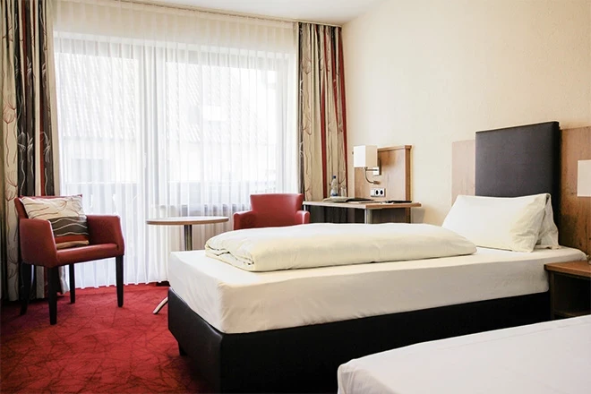 Modernes Hotelzimmer mit einem großen Bett, zwei roten Sesseln und bodenlangen Vorhängen vor einem hellen Fenster. Der Raum hat einen roten Teppich und einen dunklen Holzboden, und das Bett ist mit weißer Bettwäsche bezogen.