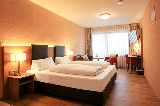 Hotelzimmer mit großem Bett, weißer Bettwäsche und gepolstertem Kopfteil. Warme, orangefarbene Beleuchtung und Vorhänge, daneben ein roter Sessel und Holzmöbel, helle Fensterfront mit Tageslicht.