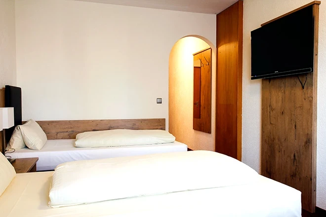 Hotelzimmer mit zwei weißen Betten, Holzmöbeln und Wandverkleidung, einem beleuchteten Rundbogen-Durchgang und einem Flachbild-Fernseher an der Wand.