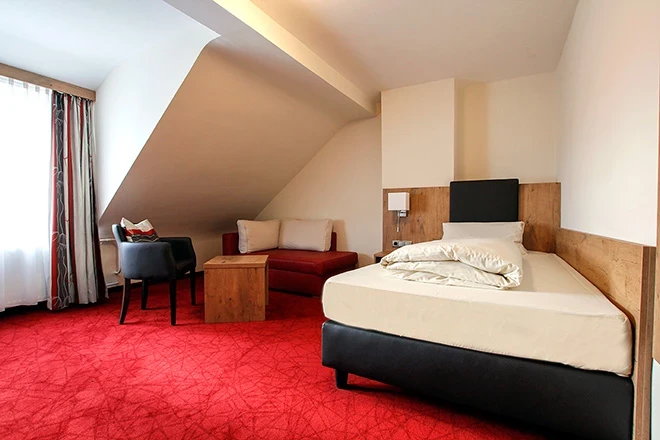 Hotelzimmer mit rotem Teppich, schräger Decke und einem Einzelbett. Holzmöbel und ein blauer Sessel komplettieren die gemütliche Einrichtung. Das Zimmer ist hell und freundlich gestaltet.