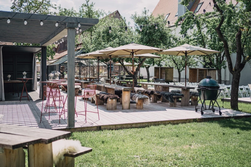 Sonnige Terrasse mit Holztischen, weißen Sonnenschirmen und Sitzgelegenheiten. Grüner Rasenbereich und ein überdachter Bereich mit roten Barhocker-Stühlen, umgeben von Bäumen und Gebäuden.