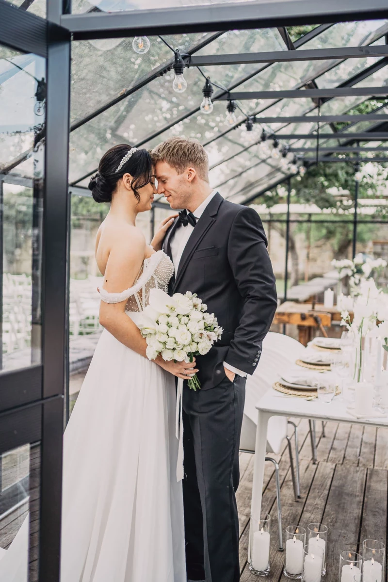 Braut und Bräutigam stehen in einem lichtdurchfluteten Glashaus, umgeben von eleganter Hochzeitsdekoration. Sie trägt ein langes weißes Kleid und hält einen weißen Blumenstrauss, während er einen schwarzen Anzug mit Fliege trägt.