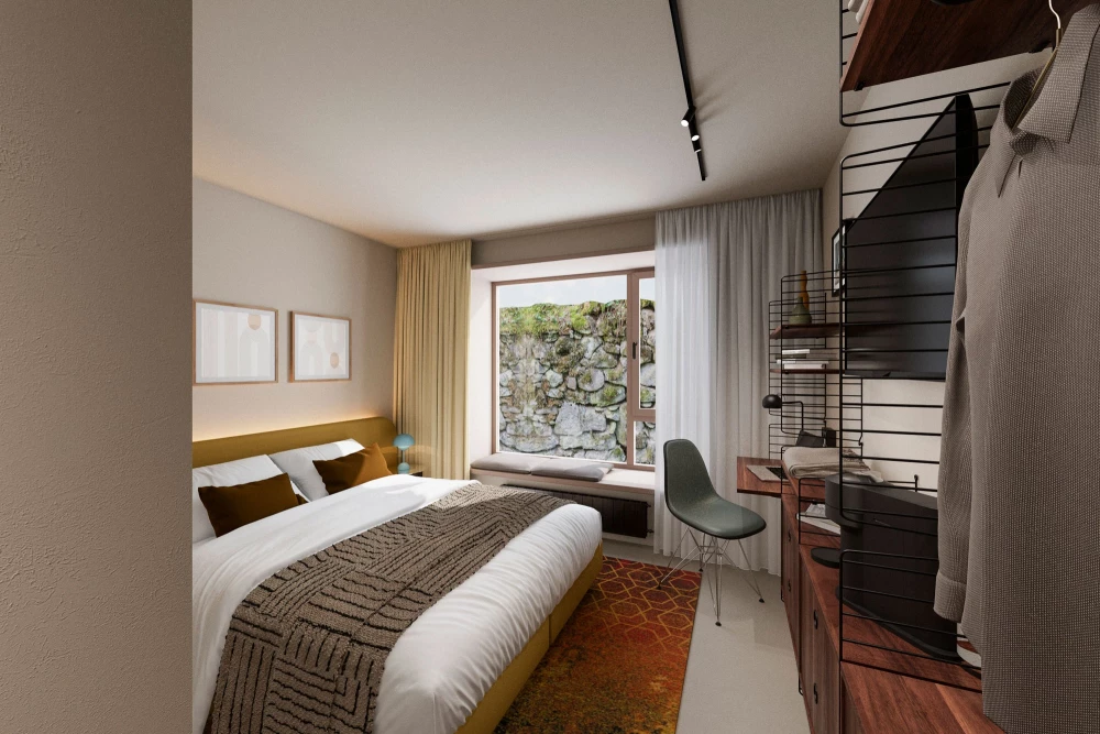 Modernes Hotelzimmer mit einem großen Bett, weißen Wänden und bodentiefen Fenster mit Blick auf eine Steinmauer. Ein orangefarbener Teppich, ein grauer Designerstuhl und ein Regal mit Büchern komplettieren das minimalistische Interieur.