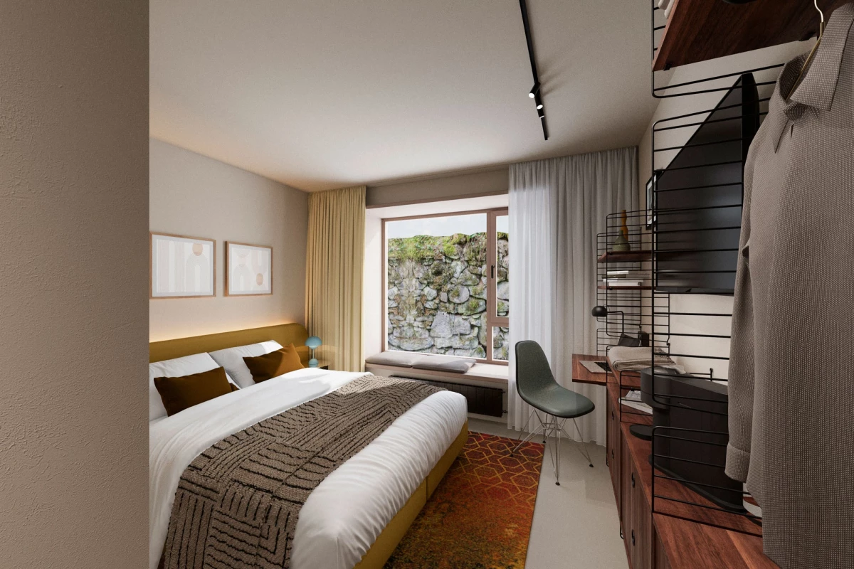 Modernes Hotelzimmer mit einem großen Bett, weißen Wänden und bodentiefen Fenster mit Blick auf eine Steinmauer. Ein orangefarbener Teppich, ein grauer Designerstuhl und ein Regal mit Büchern komplettieren das minimalistische Interieur.