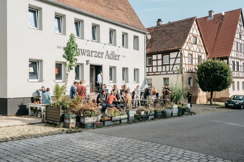 Au&szlig;enansicht des "Schwarzen Adlers" mit G&auml;sten, die vor dem wei&szlig; get&uuml;nchten Geb&auml;ude mit Fachwerkh&auml;usern im Hintergrund sitzen und stehen, vor einer mit Pflanzen und Blumen geschm&uuml;ckten Terrasse in einer malerischen deutschen Kleinstadt.