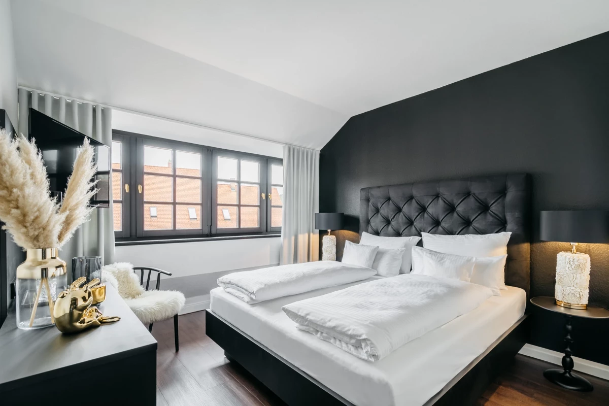 Modernes Hotelzimmer mit grauer Akzentwand und gepolstertem Kopfteil. Großes Bett mit weißer Bettwäsche, Nachttischlampen und Pampasgrasdekoration am Fenster. Helle Atmosphäre mit Holzboden und großem Fenster mit Vorhängen.