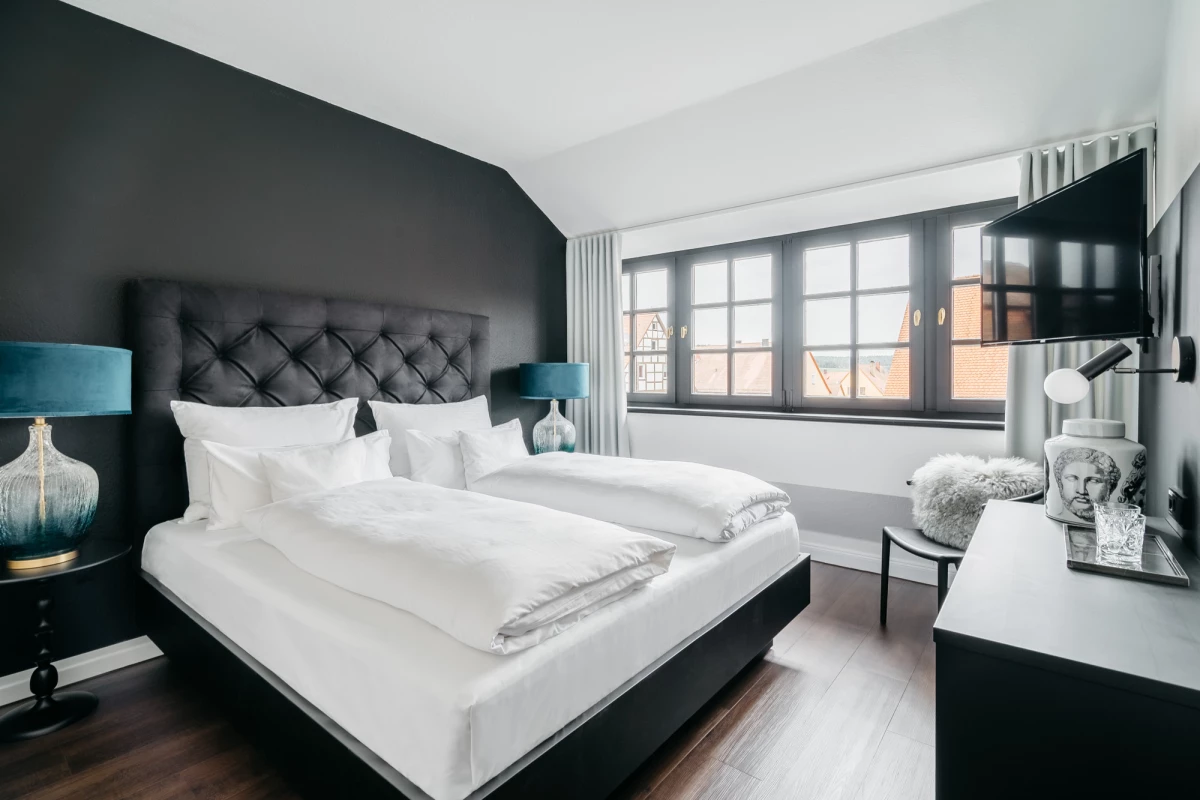Elegant eingerichtetes Hotelzimmer mit schwarzer Akzentwand, gepolstertem Kopfteil und weißer Bettwäsche. Große Fensterfront lässt natürliches Licht herein, neben dem Bett stehen blaue Designerlampen auf Nachttischen. Modernes Interieur mit dunklem Holzboden und TV an der Wand.