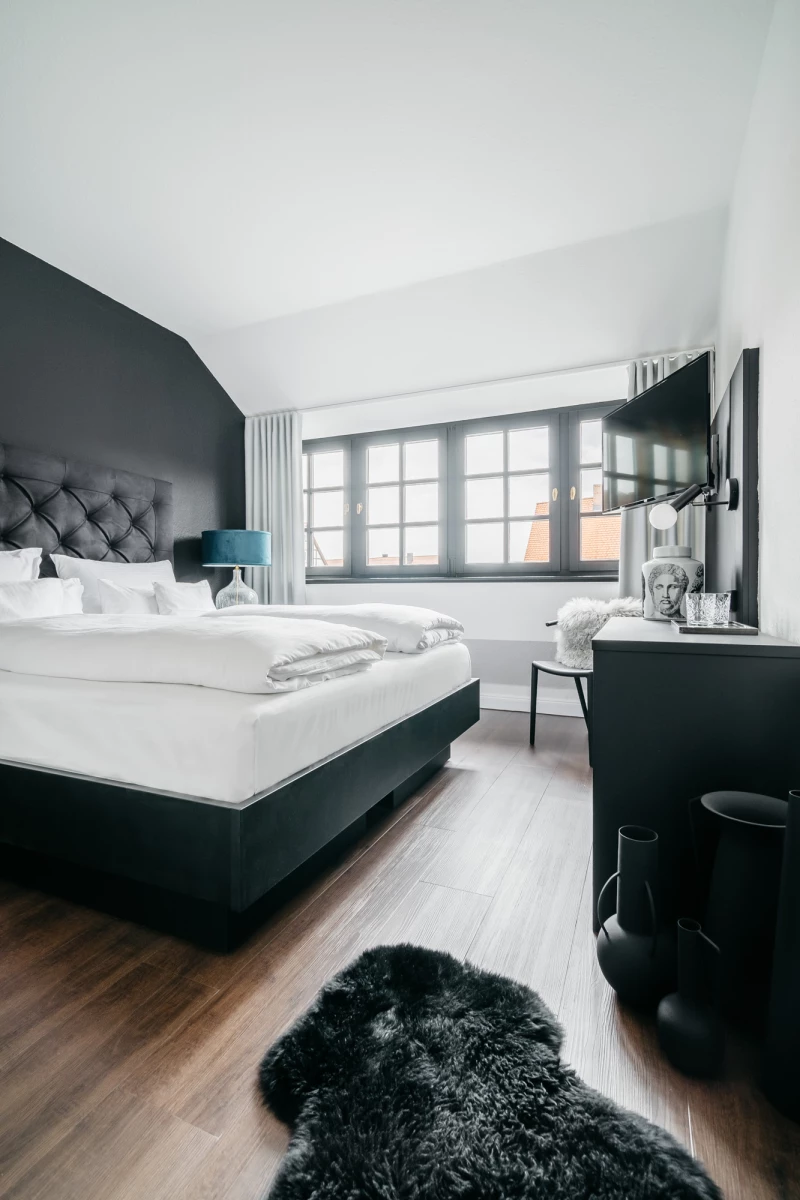 Geräumiges Hotelzimmer mit großem gepolstertem Bett in Anthrazit, hellen Bettwäschekissen und Holzboden. Großes Fenster mit Vorhängen, Flachbild-TV an der Wand und ein schwarzer Fellteppich im Vordergrund. Modernes, minimalistisches Designambiente in gedämpften Grautönen.