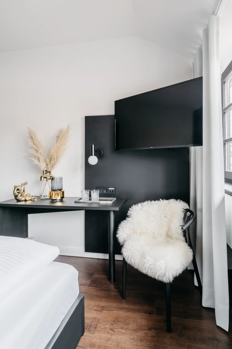 Modernes Hotelzimmer mit schwarzem Schreibtisch und weißem Fell auf einem Stuhl. Pampas-Gras in einer Vase und goldene Dekoartikel zieren den Raum, der helle Wände und einen Holzboden hat.