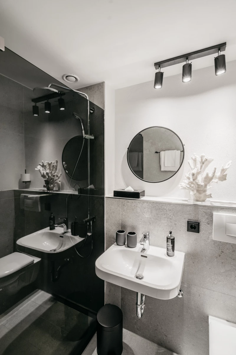 Modernes Badezimmer in Grau- und Weißtönen mit zwei Waschbecken, runden Spiegeln und minimalistischer Beleuchtung. Schwarze Armaturen und Accessoires kontrastieren mit den hellen Wänden und verleihen dem Raum einen eleganten, zeitgenössischen Charakter.