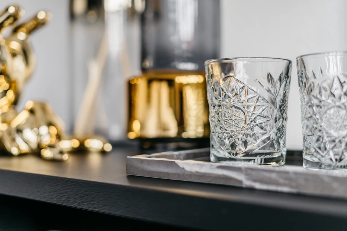 Zwei geschliffene Whiskygläser auf einem dunklen Tablett mit verschwommenen goldenen Dekorationselementen im Hintergrund. Die Gläser haben ein aufwändiges, geometrisches Schliffmuster und stehen bereit für einen eleganten Drink.