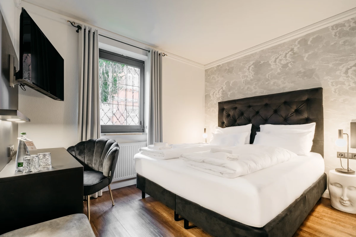 Modernes Hotelzimmer mit großem gepolstertem Bett, dunkelgrauem Kopfteil und weißer Bettwäsche. Daneben ein Schreibtisch mit einem grauen Sessel, an der Wand ein Flachbild-Fernseher und ein Fenster mit Gardinen. Der Boden ist aus dunklem Holz und die Wände sind in neutralen Farbtönen gehalten.
