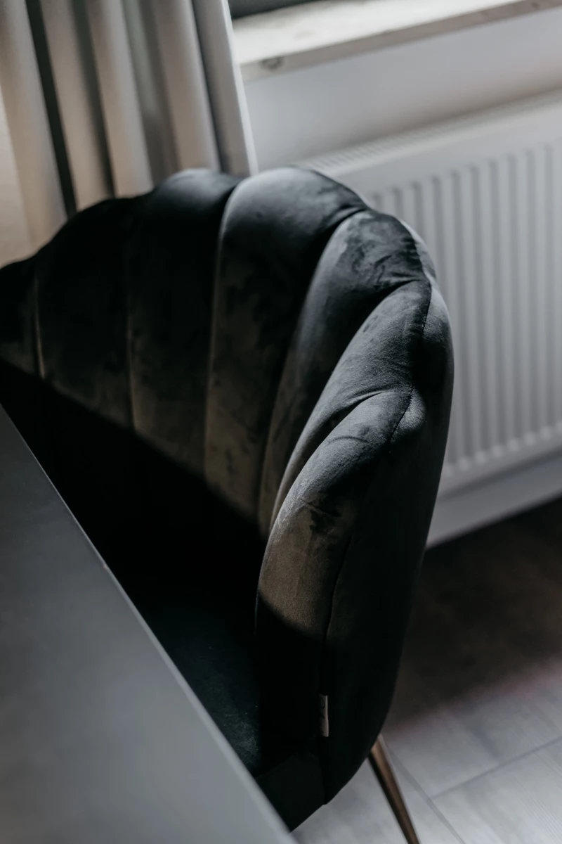 Schwarzer Sessel mit weichen Polstern an der Lehne, der vor einem weißen Heizkörper und einem Fenster mit Vorhang steht. Der Stuhl hat einen geschwungenen, eleganten Designerstil und wirkt edel und modern.