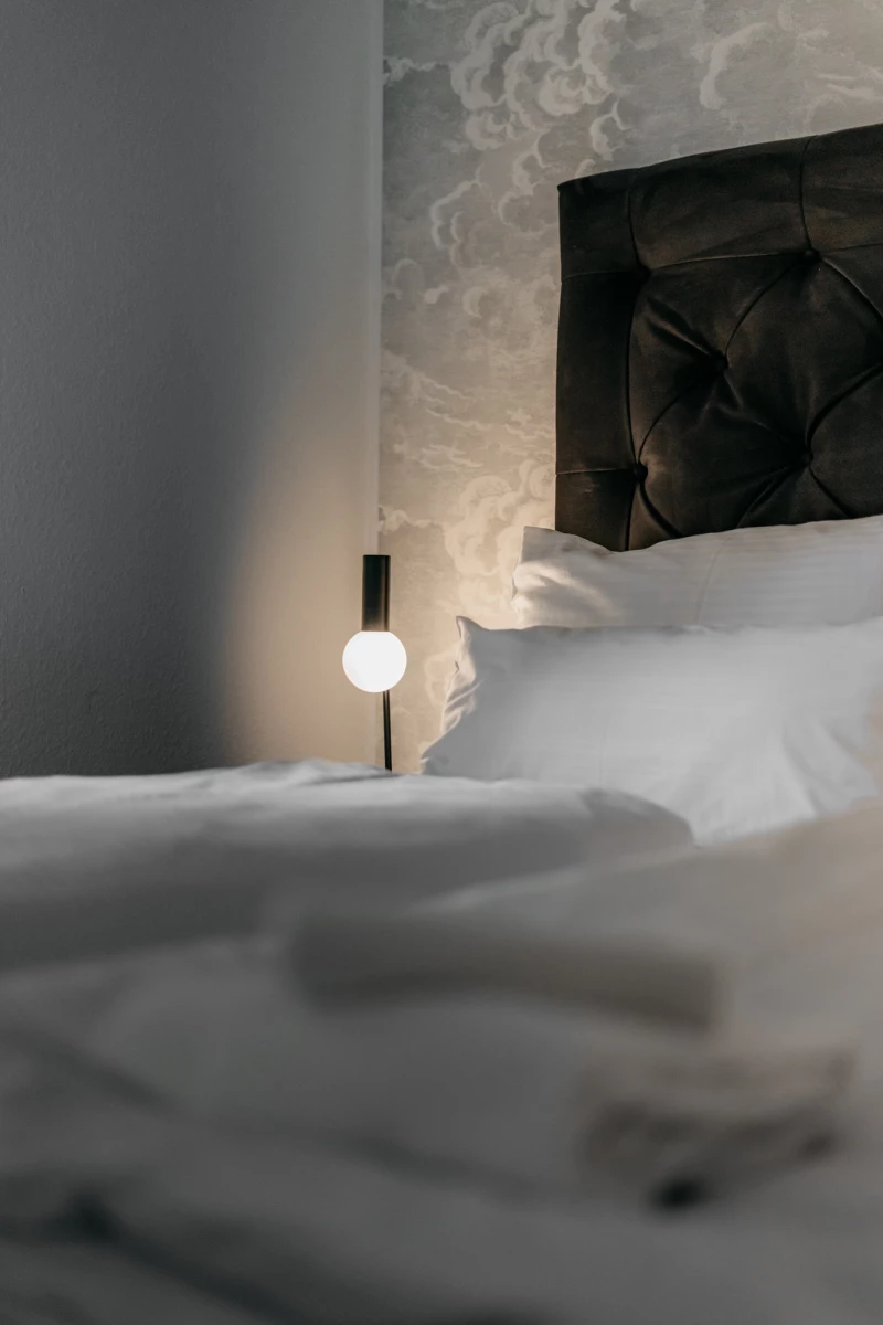 Minimalistisches Hotelzimmer mit gepolstertem Kopfteil in dunkler Farbe und weißer Bettwäsche. Eine moderne Stehlampe mit kugelförmigem Licht steht neben dem Bett, das dezent beleuchtet wird. Die Atmosphäre ist ruhig und elegant mit gedämpften Grautönen.