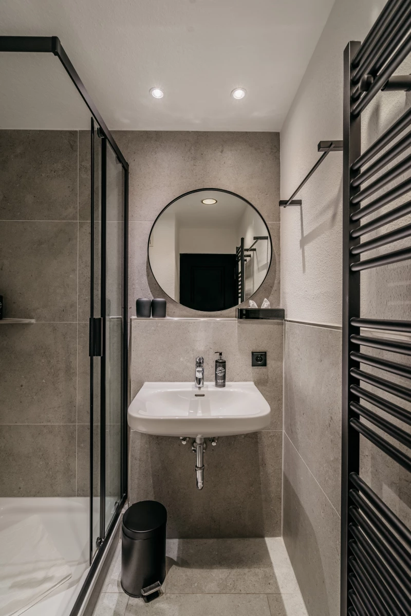 Modernes, minimalistisches Badezimmer mit grauen Wandfliesen, rundem Spiegel und weißem Waschbecken. Eine Glasdusche und ein schwarzer Papierkorb ergänzen die schlichte Einrichtung, während ein schwarzer Handtuchhalter an der Wand montiert ist.