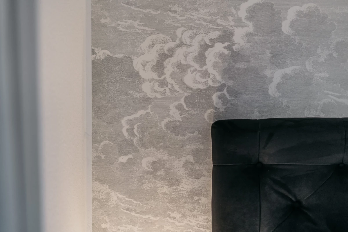 Wolkenförmige Tapete in gedämpften Grautönen mit weichen, geschwungenen Wolkenformen. Im Vordergrund ein Ausschnitt eines schwarzen gepolsterten Sitzmöbels, vermutlich Teil eines Sessels oder Sofas.