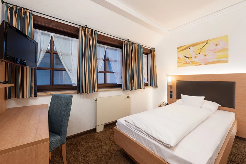 Gemütliches Einzelzimmer mit Holzmöbeln, weißem Bett, blau-beigen gestreiften Vorhängen und großen Fenstern. Auf dem Schreibtisch steht ein Flat-TV, und ein gepolsterter Stuhl steht daneben. An der Wand hängt ein gelbes Kunstwerk über dem Bett.