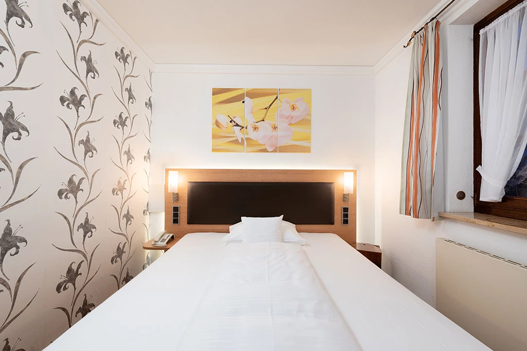 King-Size-Bett in einem hell eingerichteten Hotelzimmer mit gemusterten Tapeten, einem gelben Kunstdruck über dem Bett und gestreiften Vorhängen am Fenster. Die weiße Bettwäsche liegt ordentlich aufgeschlagen, und die Beleuchtung am Kopfteil sorgt für eine einladende Atmosphäre.