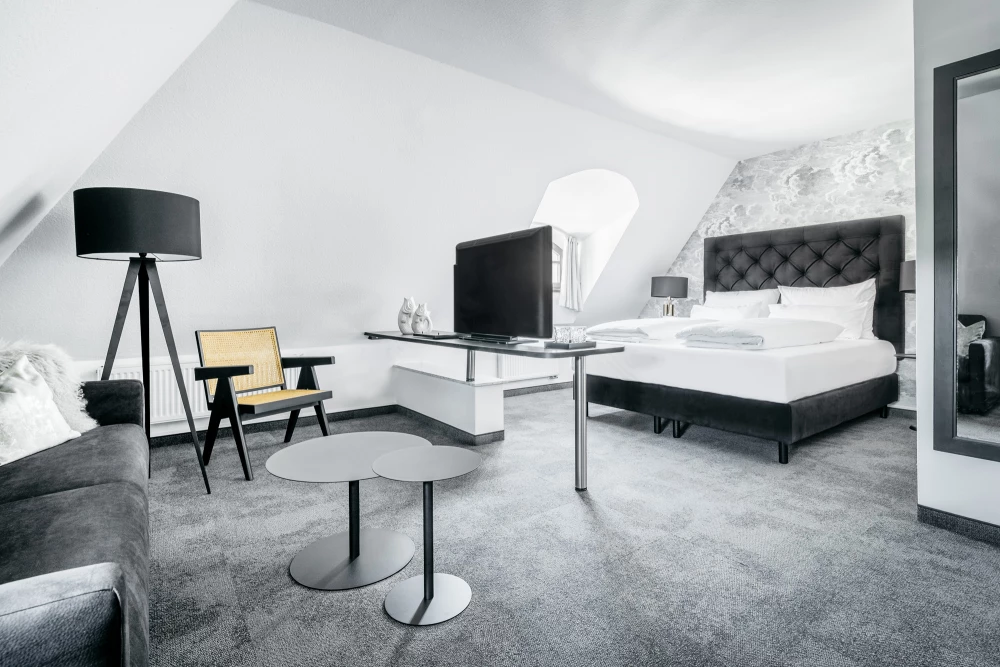 Modernes Hotelzimmer mit großem Doppelbett, gepolstertem Sessel und Stehlampe auf grauem Teppichboden. Weiße Wände und minimalistische Einrichtung in Grau- und Weißtönen, mit Flachbild-TV und eleganten Möbeln.