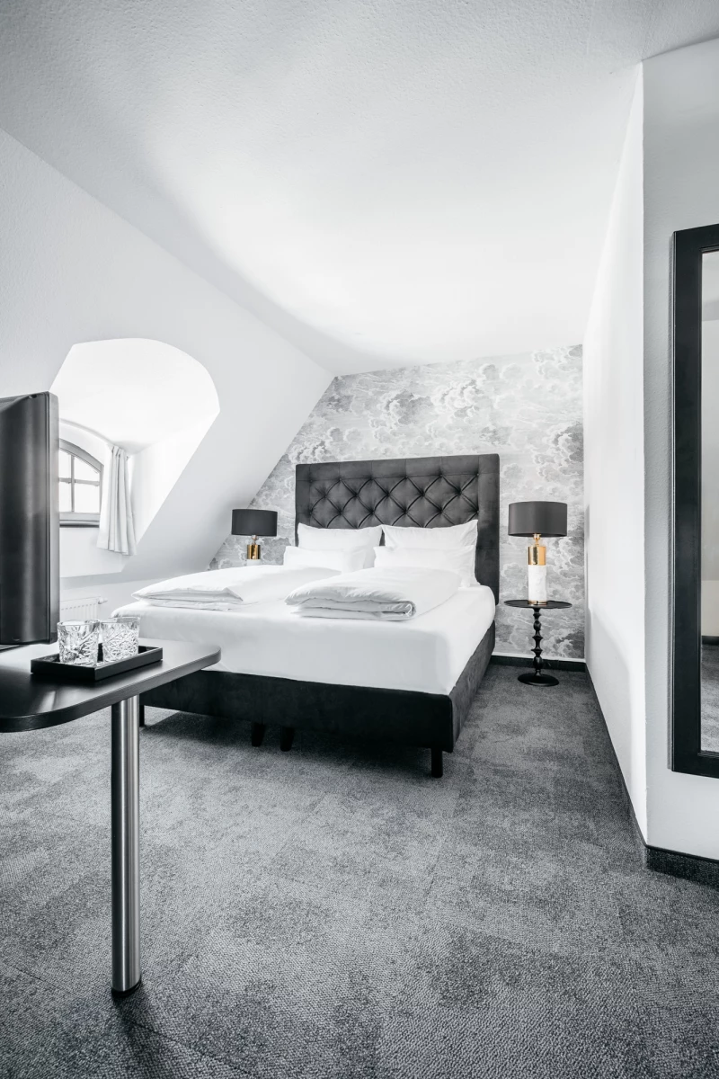 Hotelzimmer im Dachgeschoss mit einem großen grauen Bett, gepolstertem Kopfteil und schwarzen Nachttischen. Graue Teppichböden, weiße Wände und ein Dachfenster sorgen für eine helle, moderne Atmosphäre.