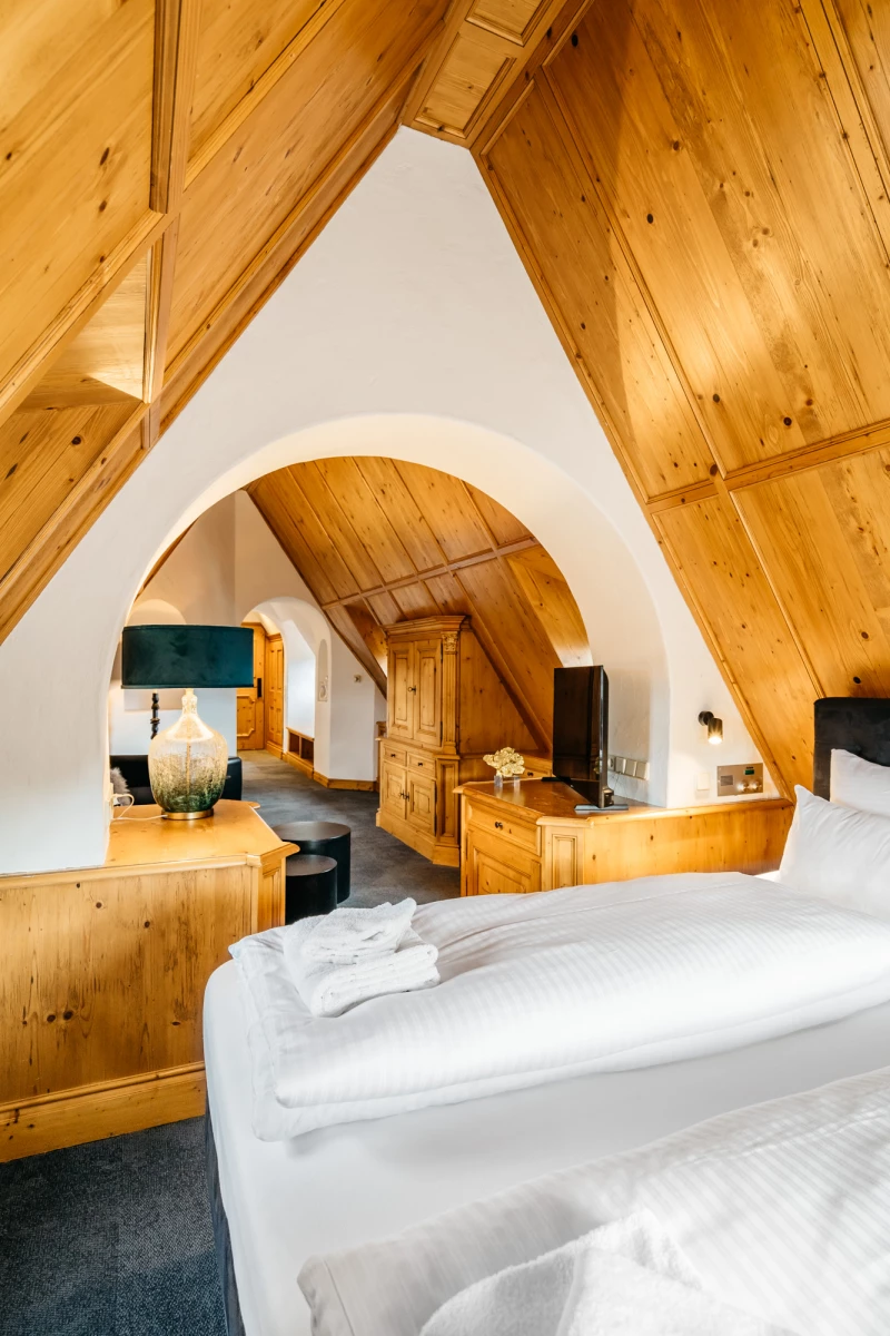 Gemütliches Dachzimmer mit Holzvertäfelungen, weiß bezogenen Betten und einem geschwungenen weißen Bogen, der verschiedene Bereiche des Raumes verbindet. Rustikale Holzmöbel und warme Holztöne schaffen eine einladende Atmosphäre unter der schrägen Dachdecke.