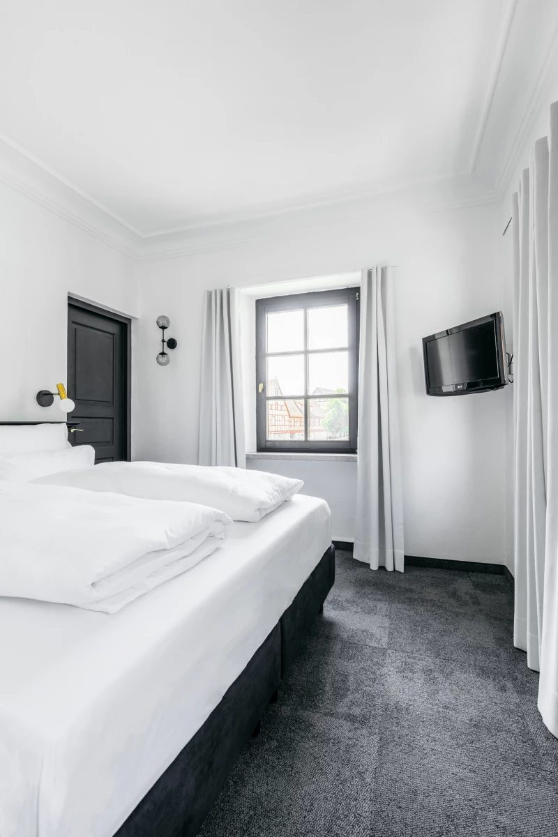 Modernes Hotelzimmer in Grau und Weiß mit einem großen weißen Bett, dunkelgrauem Teppichboden und einer Fensterfront. An der Wand hängt ein Flachbild-Fernseher, und durch das Fenster fällt gedämpftes Tageslicht.